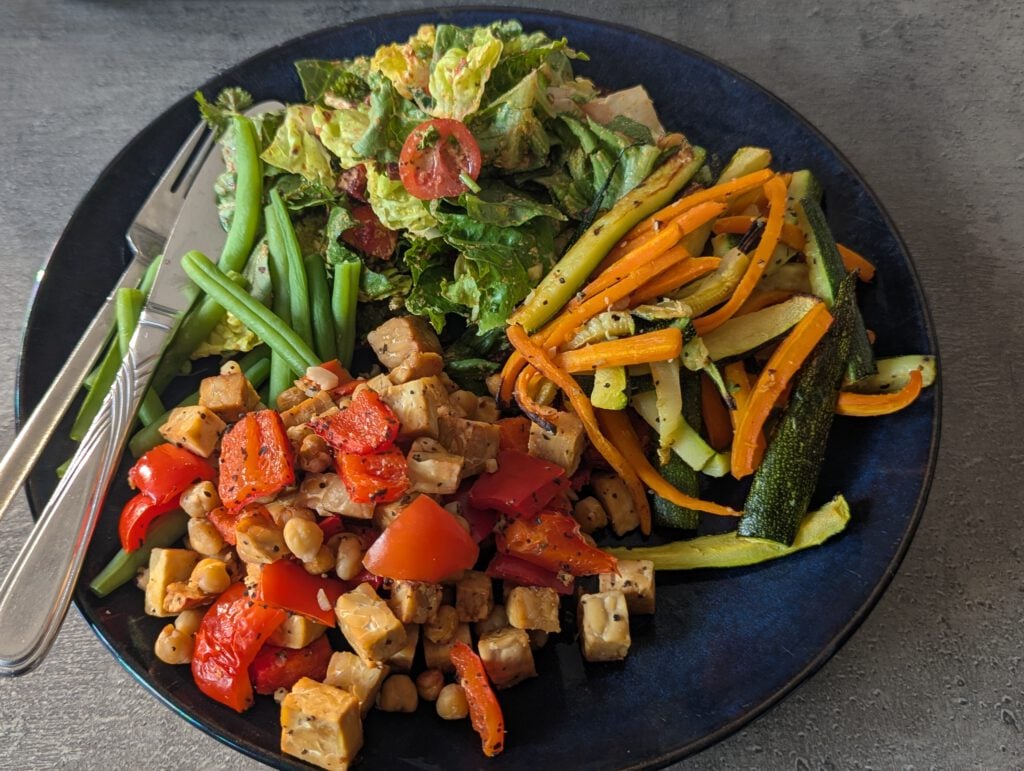 A dark blue plate of tempeh, chickpeas and roasted vegetables with salad