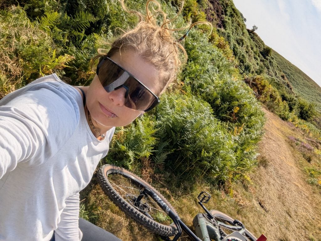 Phe Ledwell standing on a bracken-covered hillside in sunglasses, with her mountain bike beside her