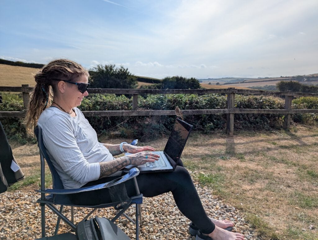 Phe Ledwell working on a laptop outdoors with fields and hedgerows behind her and a tiny bird perched on the screen